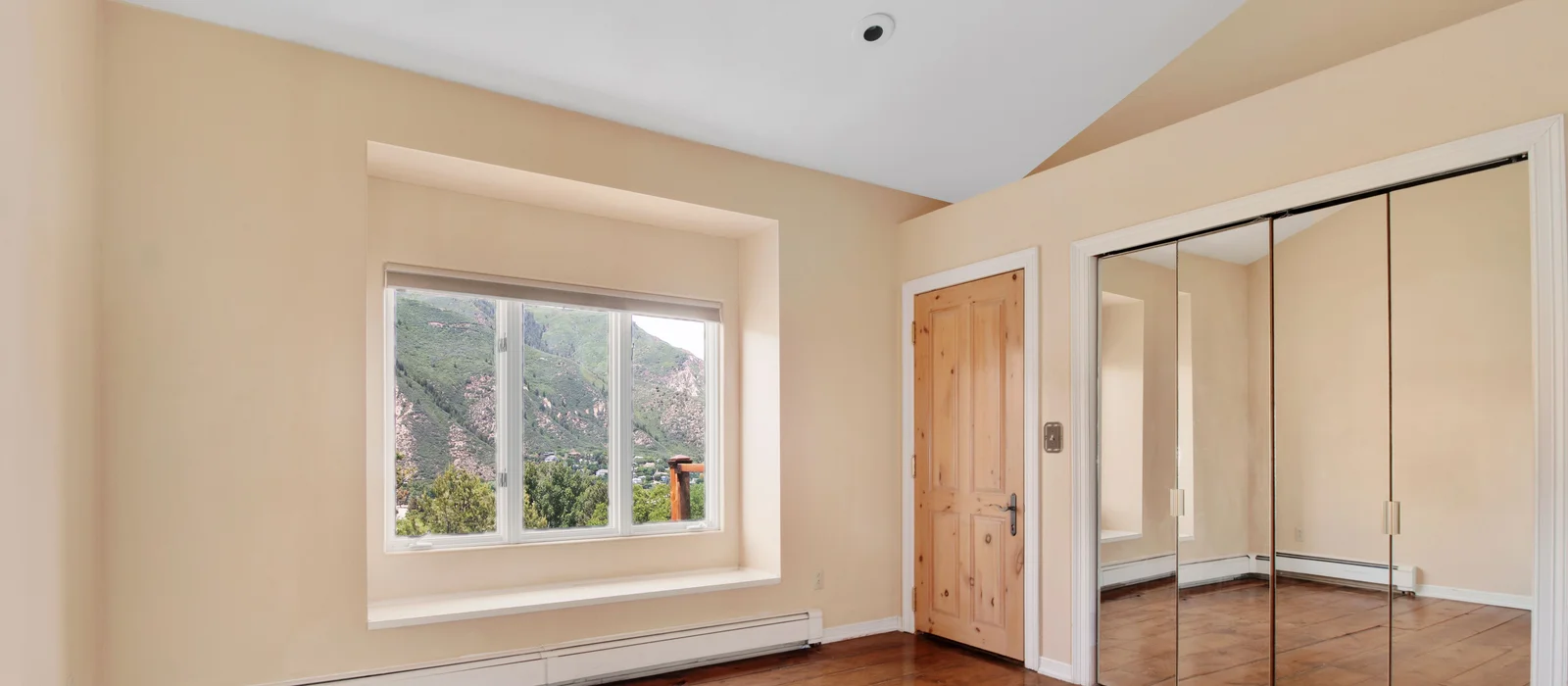 Bright bedroom with warm cream walls, large window overlooking a lush green hillside, and natural pine door with mirrored closet